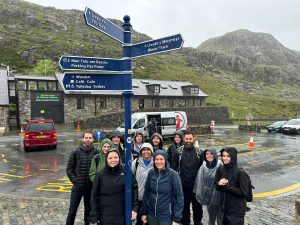 Group of people standing in the rain at the bottom of Mount Snowden in front of large sign post.